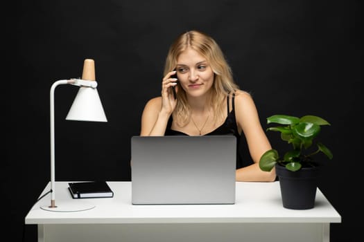 Young attractive business woman with a blond hair sitting at the table and working on a laptop and talking with a client on the phone. Young cheerful student girl talking with a smartphone. Studying