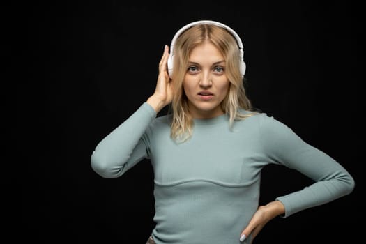 Beautiful attractive young blond woman wearing blue t-shirt and glasses in white headphones listening music and smiling on black background in studio. Relaxing and enjoying. Lifestyle