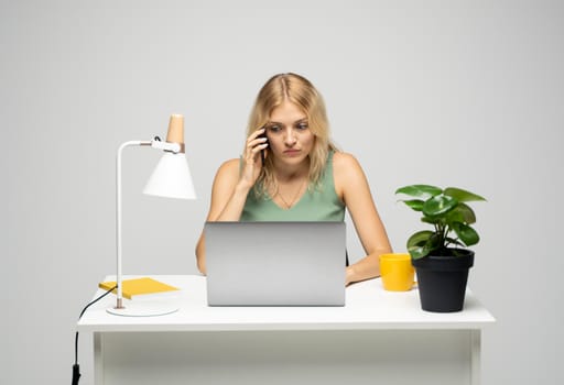 Young attractive business woman with a blond hair sitting at the table and working on a laptop and talking with a client on the phone. Young cheerful student girl talking with a smartphone. Studying