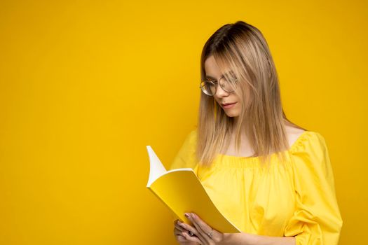 Portrait of a young student woman in a yellow t-shirt and glasses reading a book isolated over yellow background. Girl reading novel. Studying