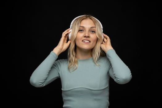 Beautiful attractive young blond woman wearing blue t-shirt and glasses in white headphones listening music and smiling on black background in studio. Relaxing and enjoying. Lifestyle