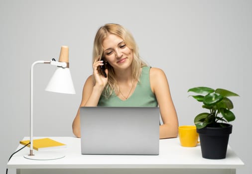 Young attractive business woman with a blond hair sitting at the table and working on a laptop and talking with a client on the phone. Young cheerful student girl talking with a smartphone. Studying