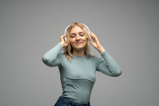 Beautiful attractive young blond woman wearing blue t-shirt and glasses in white headphones listening music and smiling on grey background in studio. Relaxing and enjoying. Lifestyle