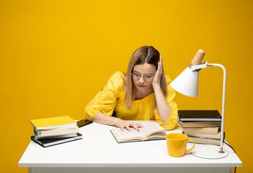 Tired young student woman in yellow casual clothes sitting at the table reading the book in library of university or college. Sitting and reading on yellow background. Studying