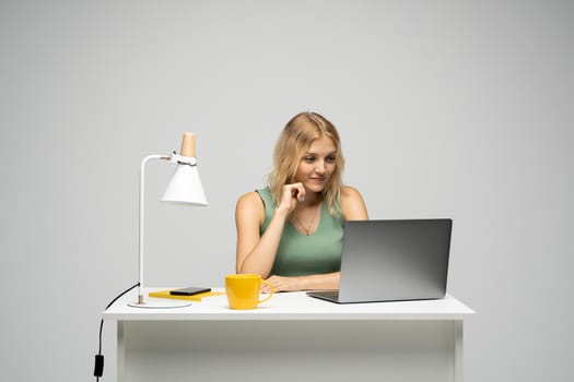 Smiling business woman working with a laptop isolated on a grey background. Portrait of a pretty young woman studying while sitting at the table with grey laptop computer, notebook.