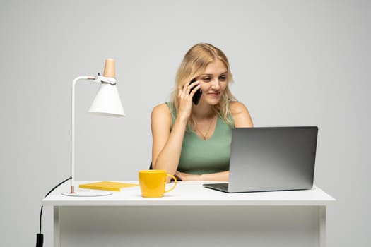 Young attractive business woman with a blond hair sitting at the table and working on a laptop and talking with a client on the phone. Young cheerful student girl talking with a smartphone. Studying