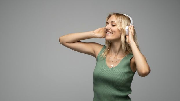 Beautiful attractive young blond woman wearing green t-shirt and glasses in white headphones listening music and smiling on grey background in studio. Relaxing and enjoying. Lifestyle