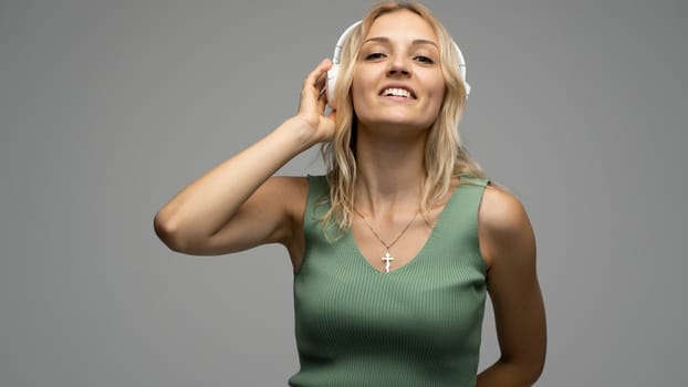 Beautiful attractive young blond woman wearing green t-shirt and glasses in white headphones listening music and smiling on grey background in studio. Relaxing and enjoying. Lifestyle