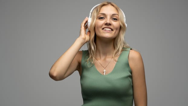 Beautiful attractive young blond woman wearing green t-shirt and glasses in white headphones listening music and smiling on grey background in studio. Relaxing and enjoying. Lifestyle