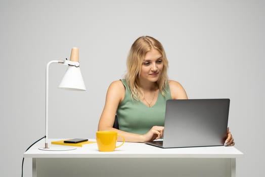 Smiling business woman working with a laptop isolated on a grey background. Portrait of a pretty young woman studying while sitting at the table with grey laptop computer, notebook.