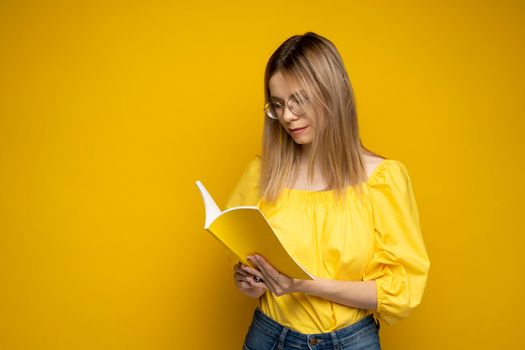 Beautiful smart young girl holding and reading book isolated on the yellow background. Portrait of attractive woman in a yellow blouse and wearing glasses reading book. Education, studying, knowledge