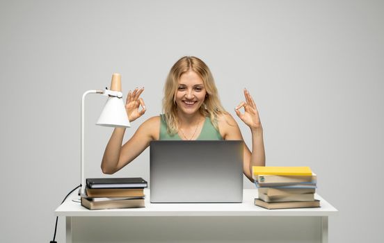 Student girl showing ok gesture with both hands, happy with completed work, smiling and looking at the camera while working on laptop computer sitting in front of big piles of books