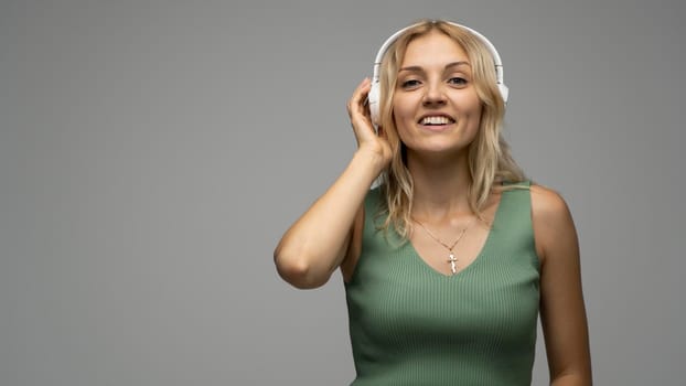 Beautiful attractive young blond woman wearing green t-shirt and glasses in white headphones listening music and smiling on grey background in studio. Relaxing and enjoying. Lifestyle