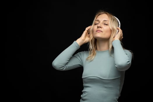 Beautiful attractive young blond woman wearing blue t-shirt and glasses in white headphones listening music and smiling on black background in studio. Relaxing and enjoying. Lifestyle