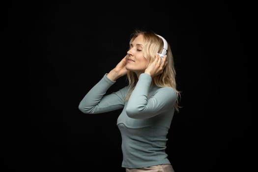 Beautiful attractive young blond woman wearing blue t-shirt and glasses in white headphones listening music and smiling on black background in studio. Relaxing and enjoying. Lifestyle