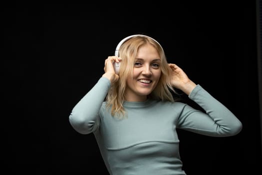 Beautiful attractive young blond woman wearing blue t-shirt and glasses in white headphones listening music and smiling on black background in studio. Relaxing and enjoying. Lifestyle