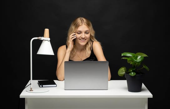 Young attractive business woman with a blond hair sitting at the table and working on a laptop and talking with a client on the phone. Young cheerful student girl talking with a smartphone. Studying