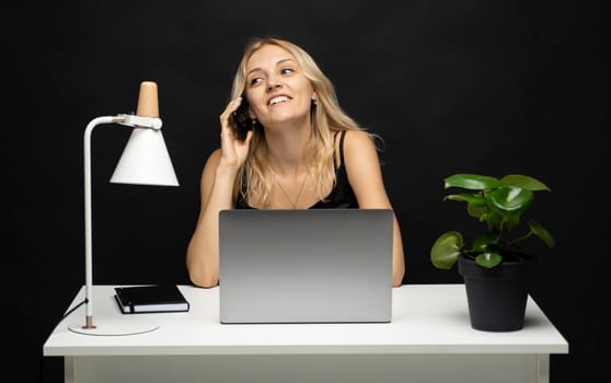 Young attractive business woman with a blond hair sitting at the table and working on a laptop and talking with a client on the phone. Young cheerful student girl talking with a smartphone. Studying