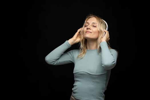 Beautiful attractive young blond woman wearing blue t-shirt and glasses in white headphones listening music and smiling on black background in studio. Relaxing and enjoying. Lifestyle