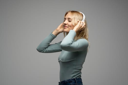 Beautiful attractive young blond woman wearing blue t-shirt and glasses in white headphones listening music and smiling on grey background in studio. Relaxing and enjoying. Lifestyle