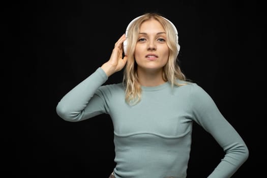 Beautiful attractive young blond woman wearing blue t-shirt and glasses in white headphones listening music and smiling on black background in studio. Relaxing and enjoying. Lifestyle