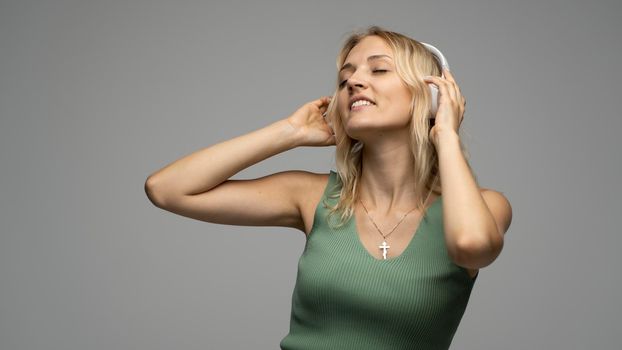 Beautiful attractive young blond woman wearing green t-shirt and glasses in white headphones listening music and smiling on grey background in studio. Relaxing and enjoying. Lifestyle