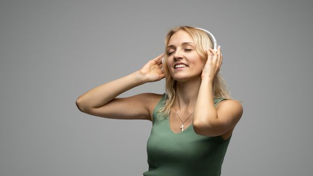 Beautiful attractive young blond woman wearing green t-shirt and glasses in white headphones listening music and smiling on grey background in studio. Relaxing and enjoying. Lifestyle