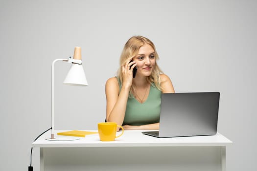 Young attractive business woman with a blond hair sitting at the table and working on a laptop and talking with a client on the phone. Young cheerful student girl talking with a smartphone. Studying