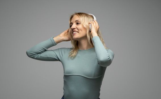 Beautiful attractive young blond woman wearing blue t-shirt and glasses in white headphones listening music and smiling on grey background in studio. Relaxing and enjoying. Lifestyle