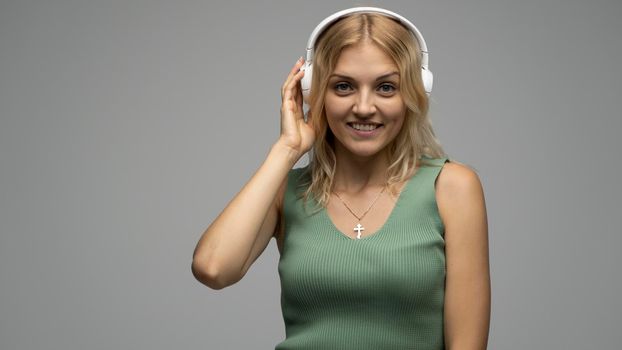 Beautiful attractive young blond woman wearing green t-shirt and glasses in white headphones listening music and smiling on grey background in studio. Relaxing and enjoying. Lifestyle