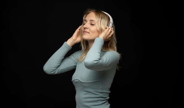 Beautiful attractive young blond woman wearing blue t-shirt and glasses in white headphones listening music and smiling on black background in studio. Relaxing and enjoying. Lifestyle