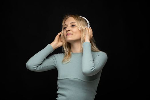 Beautiful attractive young blond woman wearing blue t-shirt and glasses in white headphones listening music and smiling on black background in studio. Relaxing and enjoying. Lifestyle