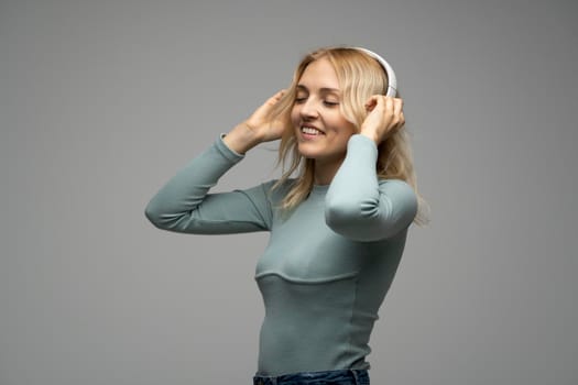 Beautiful attractive young blond woman wearing blue t-shirt and glasses in white headphones listening music and smiling on grey background in studio. Relaxing and enjoying. Lifestyle