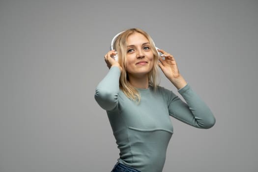 Beautiful attractive young blond woman wearing blue t-shirt and glasses in white headphones listening music and smiling on grey background in studio. Relaxing and enjoying. Lifestyle