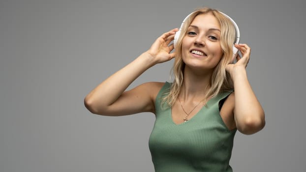 Beautiful attractive young blond woman wearing green t-shirt and glasses in white headphones listening music and smiling on grey background in studio. Relaxing and enjoying. Lifestyle