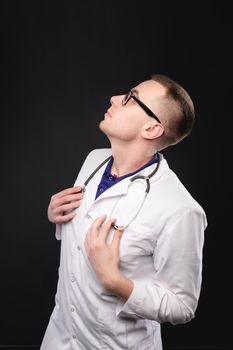 Young caucasian male doctor in a white coat uniform with a sietoscope around his neck looks up. Studio portrait on black background.