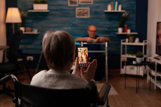 Aged person with disability using video call on smartphone for online remote communication with family. Elderly woman sitting in wheelchair at home while talking to daughter and niece