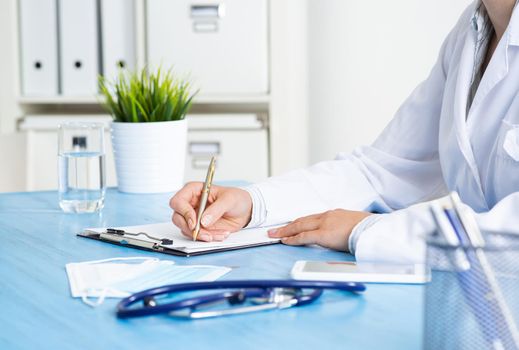 Doctor filling patient history list at clipboard. Physician in medical uniform working at desk in office. Examination and diagnosis in clinic. Doctor workplace with green plant in pot and stethoscope.