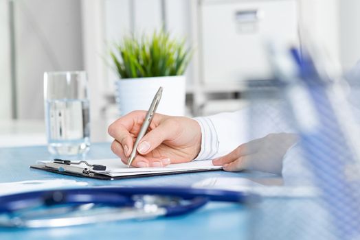 Close-up of female doctor filling medical form at clipboard. Therapist working at desk in office. Examination and diagnosis in modern clinic. Doctor workplace with green plant in pot and stethoscope.