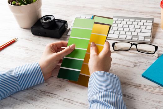 Interior designer holding color swatches at wooden desk. Office workplace with computer keyboard and camera. Brainstorming and creativity. Selection of color palette for web or product design.