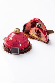Tempting dessert, whole and cut in half, of airy plum mousse on chocolate and nut shortcrust base with fruit filling topped with pink glaze and decorated with edible gold on white background