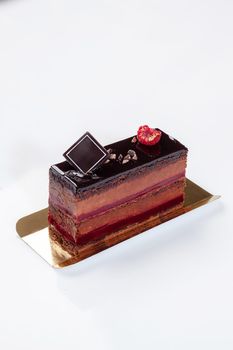 Sponge cake slice with layers of creamy cocoa mousse and jellied raspberry jam topped with chocolate icing garnished with cocoa nibs and fresh berries served on golden cardboard on white background