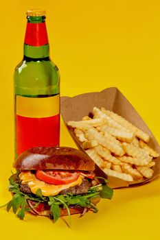 Cheeseburger with beef patty, fresh tomatoes, onion slices and greens in fluffy bun served with French fries in paper box and bottle of drink isolated on yellow background. Fast food restaurant menu
