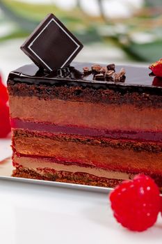 Closeup of delicious cake slice topped with cocoa glaze with layers of delicate moist chocolate sponge cake, airy chocolate mousse and raspberry confit served with fresh ripe berries