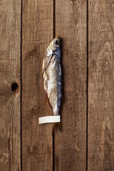 One salted air-dried Baltic herring lying on rough rustic wooden planks background, top view with copyspace. Paper label on fish tail for your logo