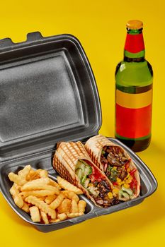 Styrofoam takeaway container with vegan burrito with mushrooms, fresh vegetables and greens, beans, corn and crispy fries on yellow background with bottle of drink. Food delivery and fast food concept