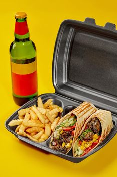 Delicious vegan burrito with mushrooms, fresh cucumber, tomato, greens, corn served in takeaway container with fries and glass bottle of drink isolated on yellow background. Food delivery concept