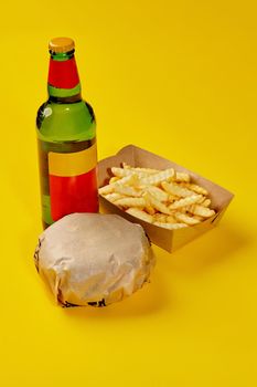 Fresh hamburger in paper wrapping, crispy French fries in carton box and glass bottle of drink isolated on yellow background. Set from fast food restaurant menu. Popular snacks concept