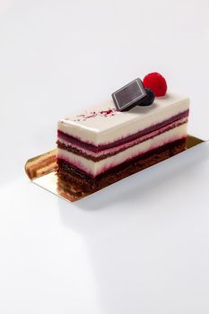 Slice of delicious raspberry dessert with layers of chocolate sponge cake, delicate cream cheese mousse and berry jelly topped with sugar icing on golden cardboard on white background