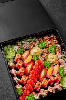 Large black cardboard box with set of delicious rolls and nigiri sushi with salmon, tuna, eel and shrimps garnished with gari, vegetables and greens. Takeaway food concept